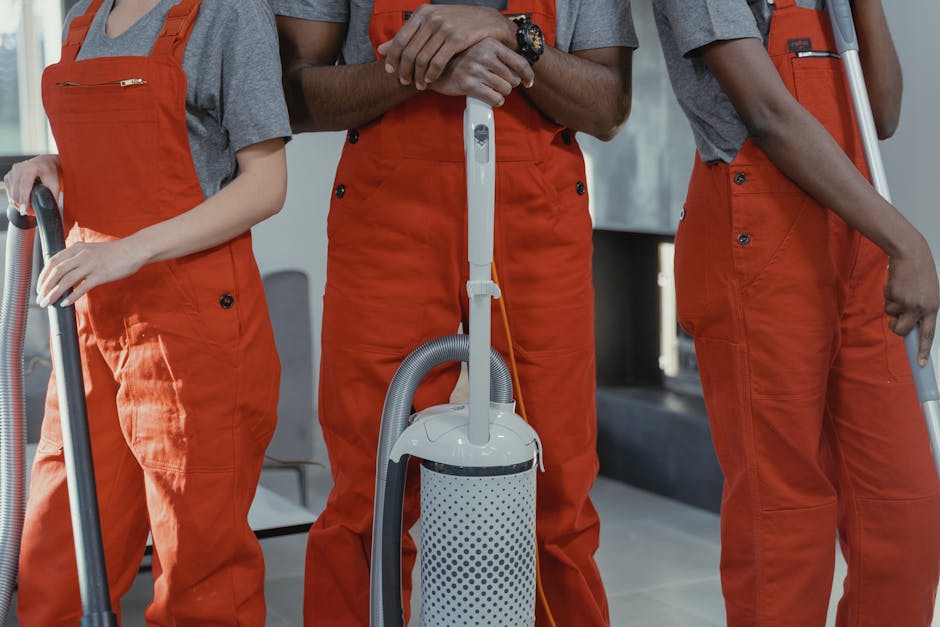 Close-up of three professional cleaners wearing orange overalls, holding vacuum cleaners and cleaning tools, standing in a modern residential space with tiled floors and a window letting in natural light. Their hands are visible, highlighting their focus on surface cleaning and deep cleaning tasks as part of a domestic cleaning service. The environment appears clean and well-maintained, emphasizing thorough sanitisation and hygiene standards. Natural light enhances the clarity of surfaces such as the tiled floor and stainless steel fixtures, showcasing their cleanliness effort, with cleaning equipment positioned prominently in the foreground. This image aligns with Tooting Cleaner’s affordable end of tenancy cleaning services in Tooting SW17.