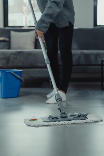 A person is engaged in surface cleaning within a modern living room, using a flat mop with a gray microfiber head to clean a smooth, light-colored tiled floor. They are wearing a gray sweater, black pants, and white socks. In the background, there is a dark gray sofa with cushions, a large window allowing natural light to brighten the space, and a blue bucket placed on the floor nearby for cleaning purposes. The room appears tidy and hygienic, with clean and shiny tile surfaces, suggesting thorough deep cleaning or sanitisation. Tooting Cleaner specialises in domestic cleaning and offers services such as end of tenancy cleaning, ensuring all surfaces, including floors and furniture, are properly cleaned and maintained for a fresh, hygienic environment.