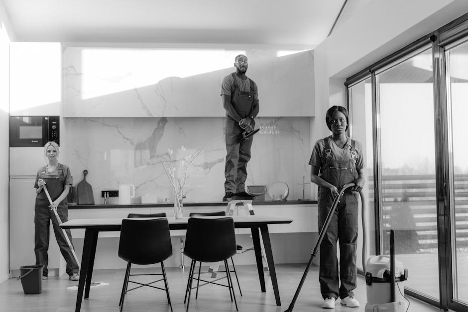 Close-up of three professional cleaners wearing orange overalls, holding vacuum cleaners and cleaning tools, standing in a modern residential space with tiled floors and a window letting in natural light. Their hands are visible, highlighting their focus on surface cleaning and deep cleaning tasks as part of a domestic cleaning service. The environment appears clean and well-maintained, emphasizing thorough sanitisation and hygiene standards. Natural light enhances the clarity of surfaces such as the tiled floor and stainless steel fixtures, showcasing their cleanliness effort, with cleaning equipment positioned prominently in the foreground. This image aligns with Tooting Cleaner’s affordable end of tenancy cleaning services in Tooting SW17.