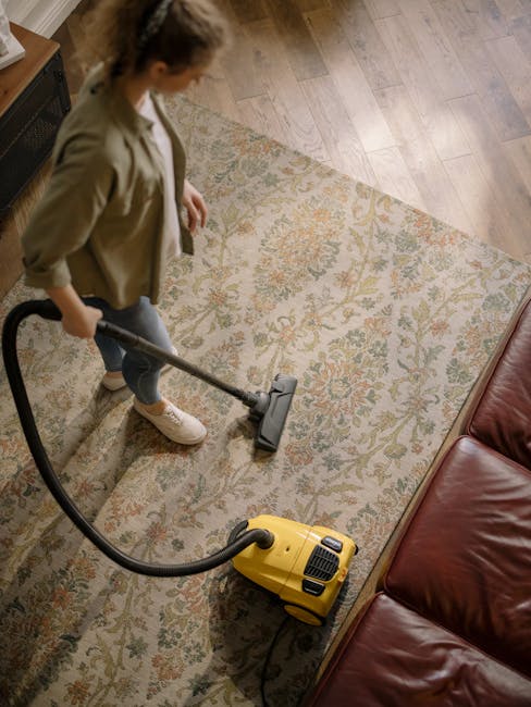 A woman is performing surface cleaning with a vacuum cleaner on a beige and floral patterned rug located in a living room. The room features light wood flooring partially visible around the edges of the rug, and a dark red leather sofa positioned against a wall. The woman is dressed in casual clothing with a beige jacket, white sneakers, and has her hair tied back. She is holding the vacuum cleaner’s handle and guiding it across the rug for deep cleaning and maintenance. The scene is well-lit, highlighting the cleanliness and freshness of the carpet, demonstrating professional domestic cleaning services offered by Tooting Cleaner in a residential setting.