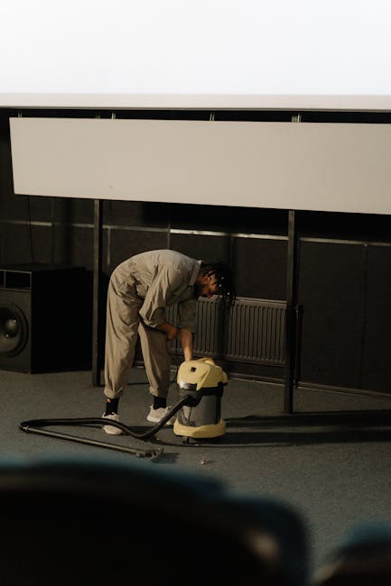 A person in a beige jumpsuit is kneeling on a carpeted floor, operating a vacuum cleaner for deep cleaning and surface sanitisation. In the background, there is a dark wall with a radiator and some electronic equipment, indicating an indoor space such as a conference room or auditorium. The vacuum cleaner is yellow and black, with a long hose, and the individual appears focused on thorough cleaning. The room has modern decor with a large white panel or screen above, and the lighting is neutral, highlighting the cleanliness of the area. This image exemplifies professional domestic or commercial cleaning practices, aligning with surface cleaning and hygiene maintenance, as emphasized by Tooting Cleaner for optimal floor and area sanitation near Tooting Bec Lido.