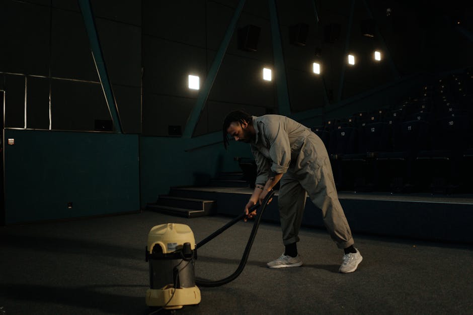 A woman is performing surface cleaning with a vacuum cleaner on a beige and floral patterned rug located in a living room. The room features light wood flooring partially visible around the edges of the rug, and a dark red leather sofa positioned against a wall. The woman is dressed in casual clothing with a beige jacket, white sneakers, and has her hair tied back. She is holding the vacuum cleaner’s handle and guiding it across the rug for deep cleaning and maintenance. The scene is well-lit, highlighting the cleanliness and freshness of the carpet, demonstrating professional domestic cleaning services offered by Tooting Cleaner in a residential setting.