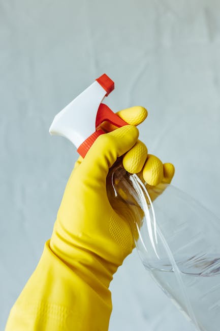 A gloved hand wearing a bright yellow rubber glove is holding a white spray bottle with a red nozzle, aiming it at a clear glass surface. The background is neutral and slightly blurred, emphasizing the cleaning action. The scene suggests surface cleaning and sanitisation, typical of professional domestic or commercial cleaning services offered by Tooting Cleaner, as highlighted in their same day deep cleaning in Tooting Broadway for urgent moves. The focus is on the cleaning tools and the act of disinfecting surfaces, ensuring a hygienic and spotless environment.