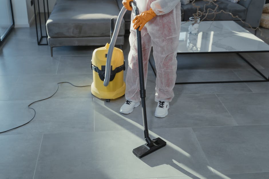 A person wearing white protective coveralls and orange rubber gloves is using a vacuum cleaner with a long hose attachment to clean a large, gray tiled floor in a modern living room. The vacuum cleaner is yellow with a black handle, positioned on the floor with its cord extending across the tiles. The surface of the floor appears spotless, reflecting light from nearby windows, indicating thorough deep cleaning. In the background, there is a gray sofa and a black metal coffee table with a glass top, and natural light illuminates the space. The scene depicts surface cleaning and maintenance as part of a professional domestic cleaning service provided by Tooting Cleaner.