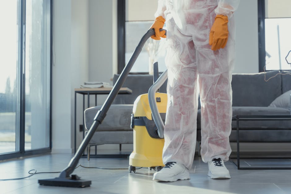 A gloved hand wearing a bright yellow rubber glove is holding a white spray bottle with a red nozzle, aiming it at a clear glass surface. The background is neutral and slightly blurred, emphasizing the cleaning action. The scene suggests surface cleaning and sanitisation, typical of professional domestic or commercial cleaning services offered by Tooting Cleaner, as highlighted in their same day deep cleaning in Tooting Broadway for urgent moves. The focus is on the cleaning tools and the act of disinfecting surfaces, ensuring a hygienic and spotless environment.